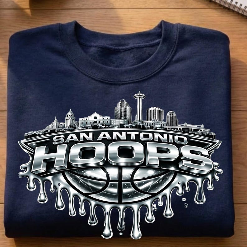 May include: Navy blue sweatshirt featuring a silver graphic of the San Antonio skyline and the words "SAN ANTONIO HOOPS" over a basketball. The design has a metallic, dripping effect, adding a dynamic visual element to the apparel.