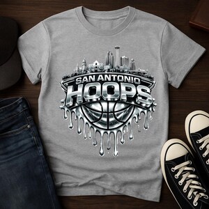 May include: A gray t-shirt featuring a San Antonio HOOPS design. The design includes a cityscape, a basketball, and the words "San Antonio HOOPS" in a metallic, dripping style. The shirt is paired with jeans and black sneakers.