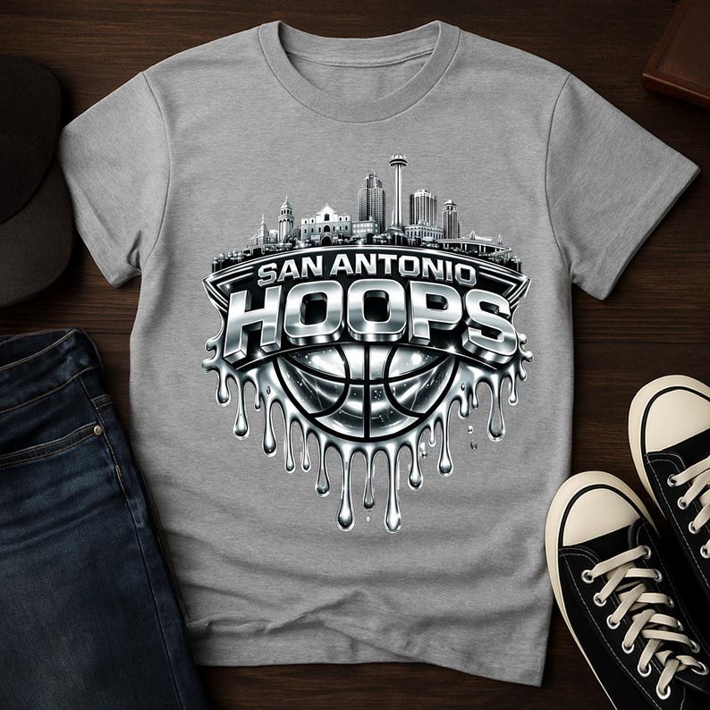 May include: A gray t-shirt featuring a San Antonio HOOPS design. The design includes a cityscape, a basketball, and the words "San Antonio HOOPS" in a metallic, dripping style. The shirt is paired with jeans and black sneakers.