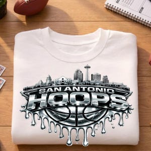 May include: White crew neck t-shirt featuring a silver and black graphic. The graphic includes the text "SAN ANTONIO HOOPS" above a basketball design and a skyline silhouette. The shirt is folded.