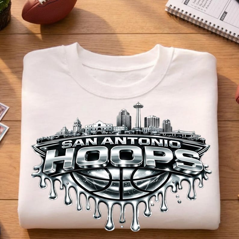May include: White crew neck t-shirt featuring a silver and black graphic. The graphic includes the text "SAN ANTONIO HOOPS" above a basketball design and a skyline silhouette. The shirt is folded.