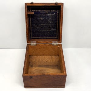 May include: Open wooden box with a hinged lid. The interior of the lid features a dark blue fabric with printed text that reads "Perfection Electro-Magnetic Machine." The box is made of a light brown wood with visible grain.