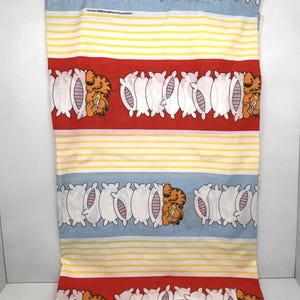 May include: A rectangular fabric pillowcase featuring a repeating pattern of the orange tabby cat Garfield resting on white pillows. The design includes horizontal bands of red, light blue, and yellow stripes with the text Garfield 1978 United Feature Syndicate Inc.