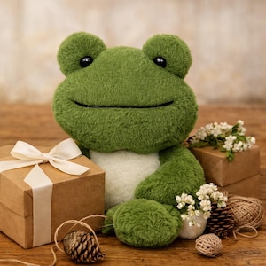 May include: A plush green frog with a white belly sits next to a wrapped gift box with a cream-colored bow. Small white flowers, pine cones, and twine are arranged around the frog, creating a whimsical scene. The frog has black eyes and a stitched smile.