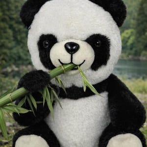 May include: A plush panda bear with black and white fur, holding a green bamboo stalk in its mouth. The panda has black eyes and ears, and is sitting in a natural outdoor setting with trees and a lake in the background.