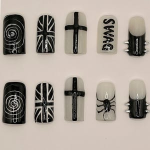 Könnte beinhalten: Ein Set aus zehn künstlichen Nägeln mit verschiedenen Designs. Die Designs umfassen eine schwarz-weiße Spirale, eine Union Jack, ein Kreuz, das Wort "SWAG", eine Spinne und Nägel mit silbernen Spitzen. Die Nägel sind quadratisch geformt.