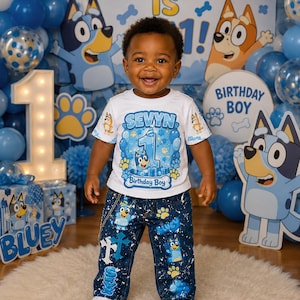 Op de afbeelding: Een kind viert zijn eerste verjaardag, gekleed in een wit T-shirt en een blauwe jeans. Het T-shirt toont de naam "SEVYN" en het cijfer "1", met Bluey-personages. De jeans is versierd met Bluey-afbeeldingen en het kind draagt witte sneakers. Blauwe en witte ballonnen zijn op de achtergrond.