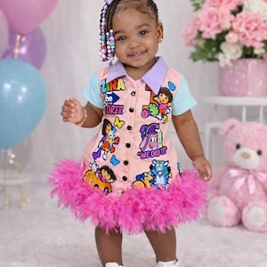 May include: A young girl wearing a pink dress with Dora the Explorer graphics. The dress has a light blue collar and sleeves, a pink feather trim, and the text "Zira the Explorer". She is wearing white sneakers.