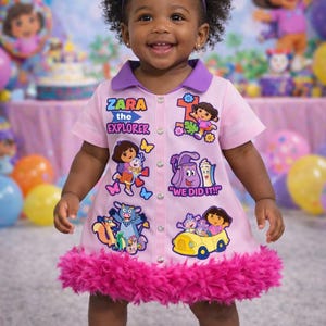 May include: A young child wearing a pink dress with Dora the Explorer graphics and the text "Zara the Explorer." The dress has a purple collar and a fluffy pink trim. The child is also wearing a colorful bow headband.