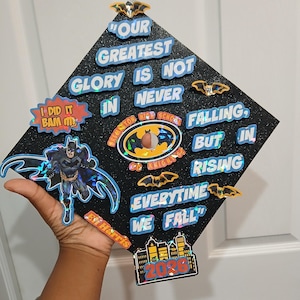 May include: A black graduation cap decorated with Batman-themed stickers and text. The cap features phrases like "Our Greatest Glory is Not Never Falling, But in Rising" and "Everytime We Fall." Includes a Batman figure and the year 2036.