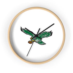 May include: A round wall clock with a light wood frame. The clock face is white and features a green and black graphic of an eagle holding a football. Black clock hands indicate the time.