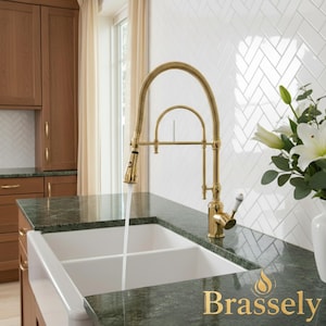 May include: A gold kitchen faucet with a pull-down sprayer is positioned over a white double sink. The faucet is set against a backdrop of white herringbone tile and a green countertop. The word "Brassely" is displayed in gold.