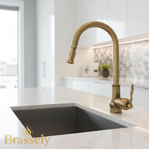 May include: A gold-coloured kitchen tap with a curved spout and pull-down sprayer is installed over a dark grey sink. The tap is set against a white countertop and white cabinet backdrop. The word "Brassely" is printed in gold at the bottom.