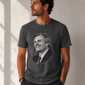 Op de afbeelding: Donkergrijs t-shirt met een zwart-wit portret van een lachende man in pak en stropdas. Het t-shirt is gemaakt van een zacht materiaal en heeft korte mouwen.