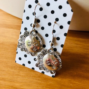 May include: Silver earrings with a sun face design. The sun face has a closed eye and a smile. The earrings are on a white card with black polka dots.