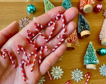 Pode incluir: Uma coleção de enfeites em forma de bengala de caramelo vermelho e branco com um laço de arame prateado para pendurar. Os enfeites estão dispostos em uma mão e uma superfície de madeira.