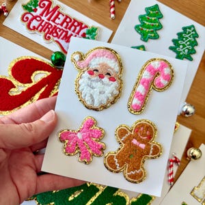 Może przedstawiać: Ręka trzyma kartkę z haftowanymi naszywkami świątecznymi, w tym Świętym Mikołajem, laską cukrową, piernikowym ludzikiem i różową kokardą. Inne karty zawierają napisy "Merry Christmas" i "HO HO HO". Naszywki mają złote obramowania.