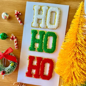 Puede incluir: Tres letras de fieltro que deletrean "HO HO" en blanco, verde y rojo con ribete de purpurina dorada. Las letras están sobre un fondo blanco con un pequeño árbol de Navidad amarillo y otras decoraciones navideñas.