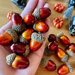 Pode incluir: Uma coleção de bolotas decorativas com acabamentos metálicos vermelhos e dourados, seguradas em uma mão. Outras decorações com tema de outono, incluindo abóboras laranja e caveiras pretas, estão espalhadas.