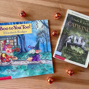Könnte beinhalten: Zwei Kinderbücher, "Boo to You Too!" und "Catwings", liegen auf einer Holzoberfläche, mit kleinen orangefarbenen Kürbisdekorationen. Das Buch "Boo to You Too!" zeigt ein Schwein im Clownkostüm und eine Hexe. "Catwings" zeigt Katzen.
