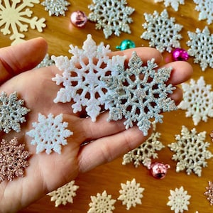 Könnte beinhalten: Eine Hand hält eine Vielzahl von glitzernden Schneeflocken-Ornamenten. Die Schneeflocken sind in verschiedenen Farben, darunter Silber, Weiß und Rosa. Die Schneeflocken haben alle einen Durchmesser von etwa 5 cm.