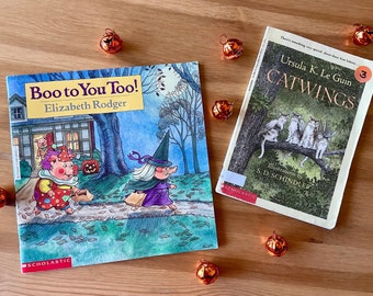 以下が含まれることがあります： 木製の表面に、「Boo to You Too！」と「Catwings」の2冊の子供向けの本が置かれ、小さなオレンジ色のカボチャの装飾が散らばっています。「Boo to You Too！」の本には、ピエロの衣装を着た豚と魔女が描かれています。「Catwings」には猫が描かれています。