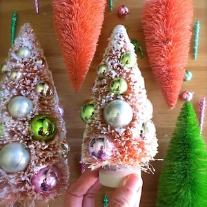 May include: Three miniature artificial Christmas trees decorated with colorful ornaments. The trees are made of brush bristles and have a white base. The trees are on a wooden surface.