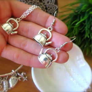 May include: Silver teapot charm necklace and earrings. The teapot charm is a detailed replica of a traditional teapot with a spout and handle. The necklace and earrings are made of silver chain.
