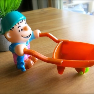 Può includere: Una colorata statuetta giocattolo di un personaggio sorridente che spinge una carriola arancione. Il personaggio indossa un cappello verde, guanti blu e una tuta rosa. La carriola ha ruote verdi. Sullo sfondo c'è una piccola pianta in vaso.