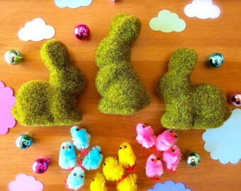 May include: Three green moss bunny figurines with pink and blue fuzzy chicks surrounding them. The bunnies are on a wooden surface with blue and white paper clouds.