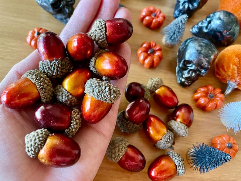 Puede incluir: Una mano sosteniendo una variedad de bellotas con tem&aacute;tica oto&ntilde;al. Las bellotas son de tonos rojos, naranjas y marrones. Algunas de las bellotas tienen un acabado brillante y met&aacute;lico.