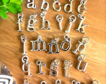 May include: A set of 26 silver letter charms, each charm is a different letter of the alphabet. The charms are arranged in alphabetical order on a wooden surface.