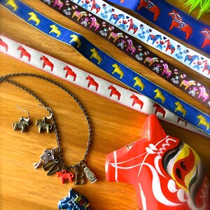 Puede incluir: Un surtido de art&iacute;culos con tem&aacute;tica de caballos Dala. Incluye cintas estampadas en azul, blanco y marr&oacute;n, un collar con colgantes, pendientes y dos caballos Dala de madera pintados en rojo y azul. Las cintas presentan motivos de caballos.