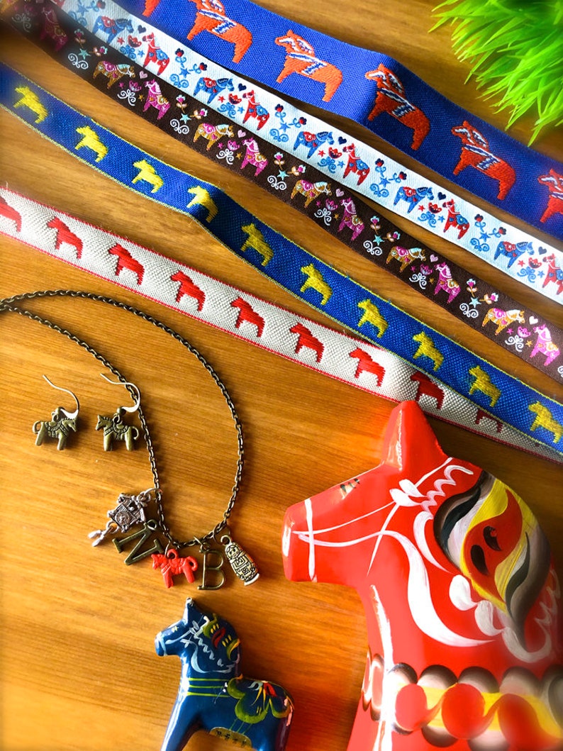 Puede incluir: Un surtido de art&iacute;culos con tem&aacute;tica de caballos Dala. Incluye cintas estampadas en azul, blanco y marr&oacute;n, un collar con colgantes, pendientes y dos caballos Dala de madera pintados en rojo y azul. Las cintas presentan motivos de caballos.