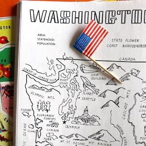 Könnte beinhalten: Eine Schwarzweißillustration einer Karte des Bundesstaates Washington mit verschiedenen Sehenswürdigkeiten und geografischen Merkmalen, die beschriftet sind. Die Karte umfasst die Olympischen Berge, Puget Sound, die Straße von Juan de Fuca, Seattle, Olympia, Mount Rainier, Mount St. Helens, den Columbia River und den Grand Coulee Dam. Eine kleine amerikanische Flagge ist auf die Karte gesteckt.