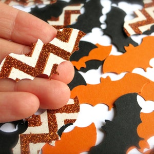 K&ouml;nnte beinhalten: Nahaufnahme einer Hand, die eine wei&szlig;e und orangefarbene Glitzer-Fledermaus mit Chevron-Muster h&auml;lt. Die Fledermaus ist von anderen Flederm&auml;usen in Orange und Schwarz umgeben.