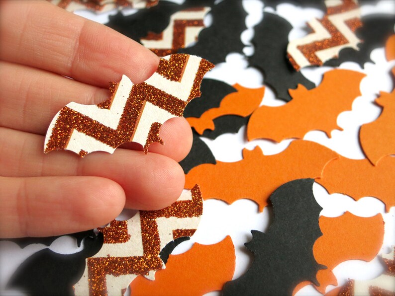 K&ouml;nnte beinhalten: Nahaufnahme einer Hand, die eine wei&szlig;e und orangefarbene Glitzer-Fledermaus mit Chevron-Muster h&auml;lt. Die Fledermaus ist von anderen Flederm&auml;usen in Orange und Schwarz umgeben.