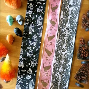 Puede incluir: Tres cintas diferentes con tem&aacute;tica de Halloween. La primera cinta es negra con dise&ntilde;os de cristales blancos y calaveras. La segunda cinta es rosa con gatos negros y calaveras. La tercera cinta es blanca con encaje negro y dise&ntilde;os de calaveras.