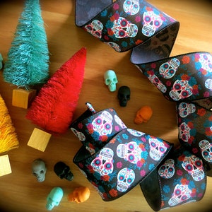 Puede incluir: Una cinta negra con un patr&oacute;n repetido de calaveras de az&uacute;car coloridas y flores. La cinta est&aacute; enroscada y se muestra sobre una superficie de madera con peque&ntilde;os &aacute;rboles de cepillo coloridos y calaveras decorativas.
