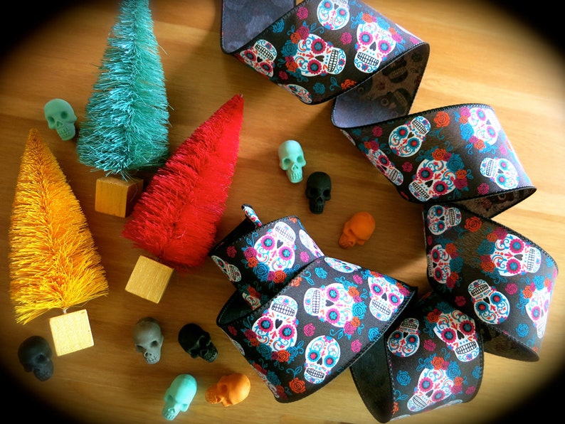 Puede incluir: Una cinta negra con un patr&oacute;n repetido de calaveras de az&uacute;car coloridas y flores. La cinta est&aacute; enroscada y se muestra sobre una superficie de madera con peque&ntilde;os &aacute;rboles de cepillo coloridos y calaveras decorativas.