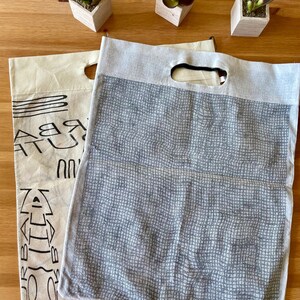 May include: Two reusable shopping bags. One is a beige canvas bag with black text and a black mesh pocket. The other is a gray mesh bag with a black handle.