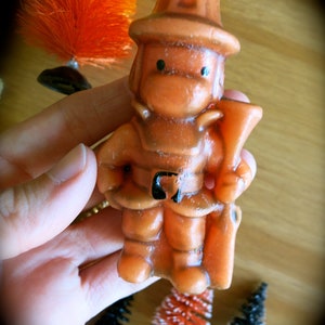 May include: Orange and brown figurine of a firefighter holding a hose. The figurine is made of wax and has a simple, cartoon-like design.