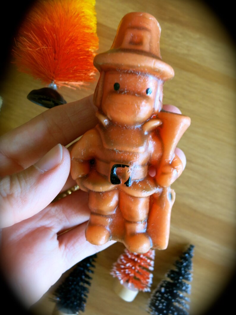 May include: Orange and brown figurine of a firefighter holding a hose. The figurine is made of wax and has a simple, cartoon-like design.