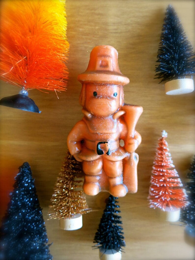 May include: A small orange, brown, and white figurine of a firefighter wearing a hat. The figurine is surrounded by small artificial Christmas trees in various colors.