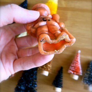 May include: An orange candle shaped like a person sitting down. The candle has a black line drawn on it. The candle is being held by a hand.