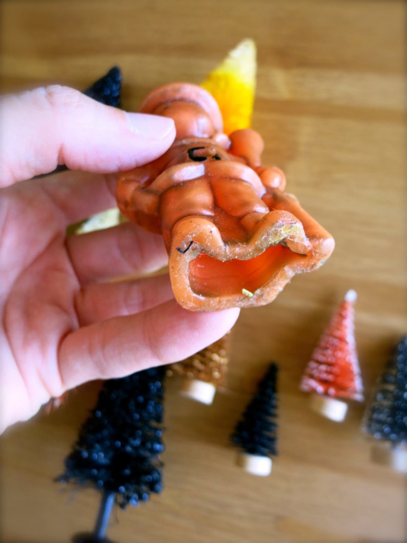 May include: An orange candle shaped like a person sitting down. The candle has a black line drawn on it. The candle is being held by a hand.