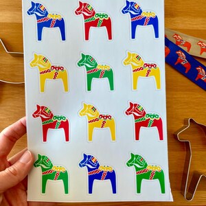 Puede incluir: Hoja de calcoman&iacute;as de caballos Dala en varios colores, incluyendo rojo, azul, amarillo y verde. La hoja tambi&eacute;n tiene el texto "Dala Horse Decals" y el n&uacute;mero "#14-138". Cintas decorativas y un cortador de galletas con forma de caballo tambi&eacute;n son visibles.