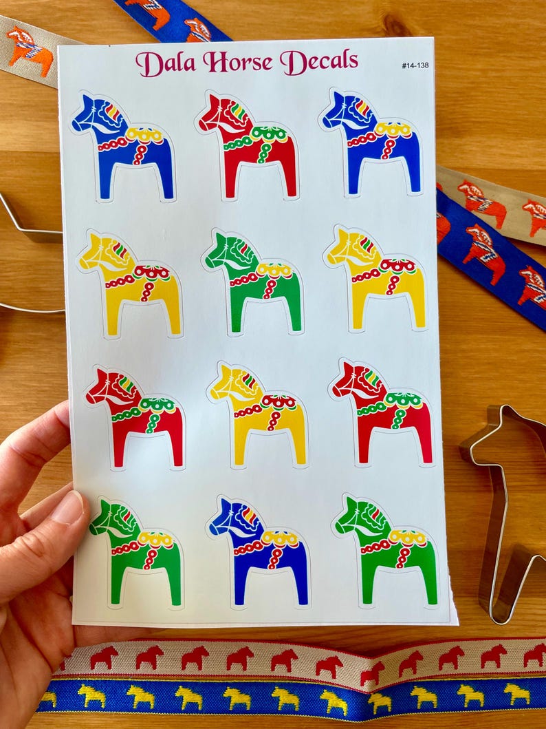 Puede incluir: Hoja de calcoman&iacute;as de caballos Dala en varios colores, incluyendo rojo, azul, amarillo y verde. La hoja tambi&eacute;n tiene el texto "Dala Horse Decals" y el n&uacute;mero "#14-138". Cintas decorativas y un cortador de galletas con forma de caballo tambi&eacute;n son visibles.
