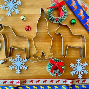 Puede incluir: Decoraciones navide&ntilde;as sobre una superficie de madera. La imagen incluye cortadores de galletas de metal con forma de caballo y gnomo, peque&ntilde;as coronas con lazos rojos, adornos de copos de nieve y cintas decorativas con motivos de caballos.