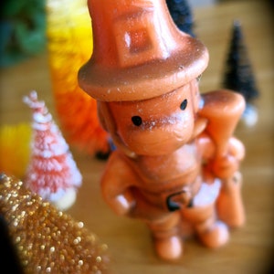 May include: A small, orange figurine of a person wearing a hat and holding a small, brown object. The figurine is made of plastic and has a whimsical, cartoonish style.